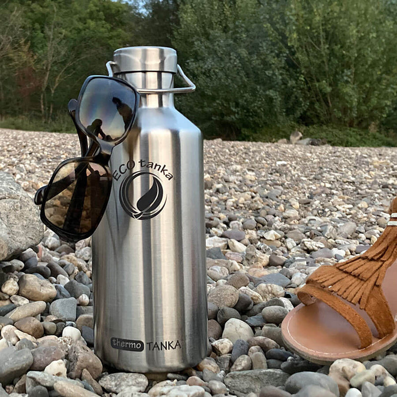 thermo Edelstahl Trinkflasche in silber 1,2l am Kiesstrand am Rhein mit Sonnenbrille und Sandale