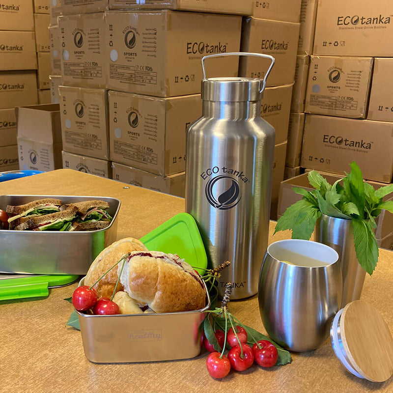 auf einem Tisch im ECOTanka Verwsandlager eine pocketBOx und eine lunchbox mit belegten Broten und Obst und eine megatNAKA mit Wasser und ein ECOtanka Cup mit Tee